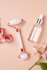 Glass bottle with oil, face roller for face massage on a beige background with flowers. Cosmetic facial skin care and spa. Natural treatment concept.