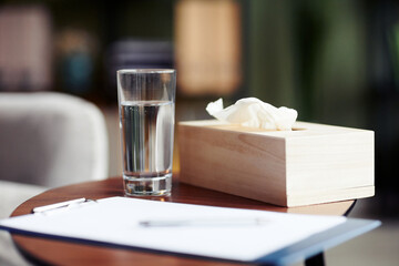 Glass of fresh water, blank sheet on clipboard and napkins for client on table in office of therapist