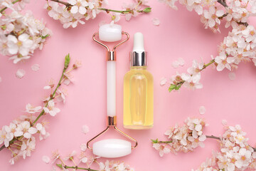 Glass bottle with oil, face roller for face massage on a pink background with blooming cherry....