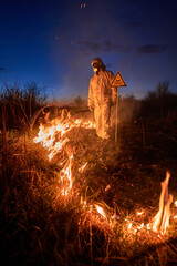 Ecologist extinguishing fire in field at night. Man in suit and gas mask near burning grass with...