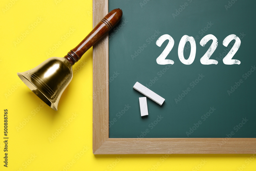 Sticker Back to school 2022. Golden bell, green board and pieces of chalk on yellow background, flat lay