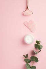Skincare concept. Top view vertical photo of rose quartz roller gua sha cream bottle and eucalyptus sprig on isolated pastel pink background