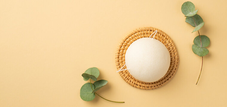 Top view photo of white konjac facial sponge on wicker stand and eucalyptus on isolated pastel beige background with copyspace