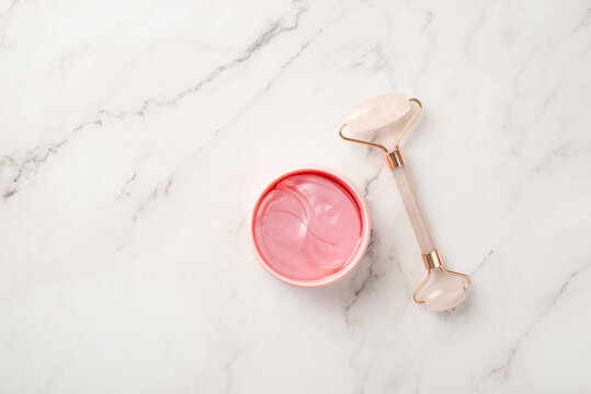 Skincare concept. Top view photo of rose quartz roller and pink eye patches on white marble background with copyspace