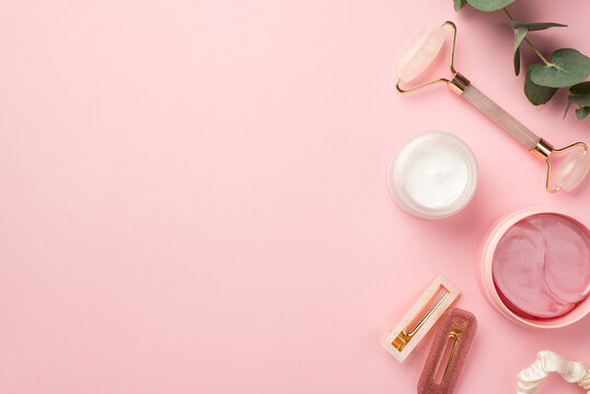 Skincare concept. Top view photo of rose quartz roller pink eye patches cream jar pink stylish barrettes scrunchy and eucalyptus on pastel pink background with blank space