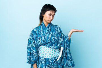 Young woman wearing kimono over isolated blue background holding copyspace with doubts
