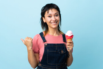 Young Uruguayan girl holding a cornet ice cream over isolated blue background pointing to the side...