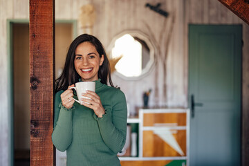 Portrait of a smiling female, leaning on the wooden pillar, enjoying her coffee.