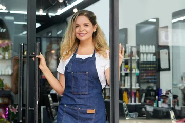 Lichtdoorlatende rolgordijnen Schoonheidssalon Positive young hairdresser in denim apron opening salon door to let clients in