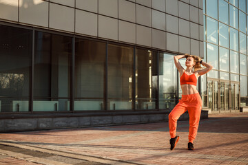 Athletic woman in orange sportswear is posing in front of the glass urban building, copy space.