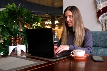 A young brunette woman is working at a laptop in a cafe. The concept of freelancing, remote work, distance learning, remote work. Freelancer. Coworking.