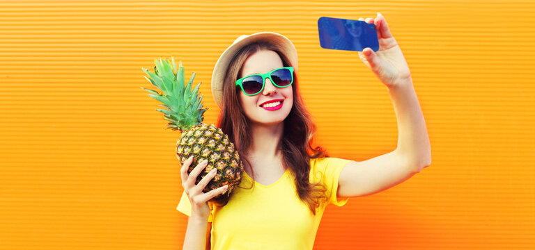 Summer portrait close up of happy smiling woman with pineapple taking selfie by smartphone wearing straw hat, sunglasses on vivid background