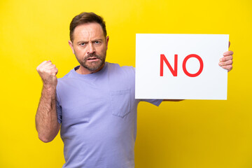 Middle age caucasian man isolated on yellow background holding a placard with text NO and angry