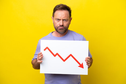 Middle age caucasian man isolated on yellow background holding a sign with a decreasing statistics arrow symbol with sad expression