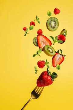 Fresh strawberries, kiwi slices and mint leaves thrown in the air, with fork, flying and levitating on a vibrant yellow background. Creative food concept. Summer fruit or healthy diet eating idea.