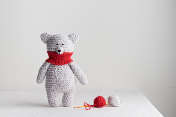 Crochet knitting cute gray, white, polar teddy bear toy with a red scarf stands on the table, homemade amigurumi with knitting hook and yarn, copy space