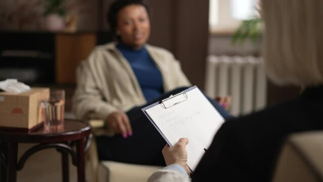 Psychologist listening to female patient and holding clipboard during consultation in office spbas. 4k Close view of gray-haired person listens to story and holds document in hand, American African