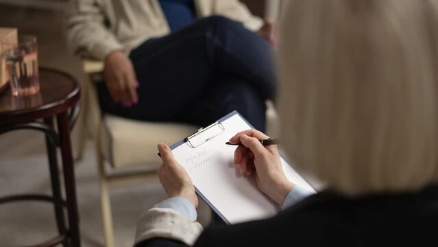 Close view of female psychologist is counseling client and writing during psychotherapy spbas. 4k Female psychotherapist listens to patient's story and holds clipboard in hand, takes notes and sits in