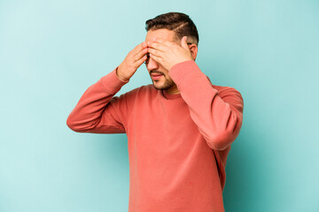 Young hispanic man isolated on blue background afraid covering eyes with hands.