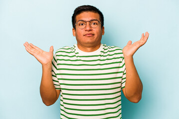 Fototapeta premium Young hispanic man isolated on blue background confused and doubtful shrugging shoulders to hold a copy space.