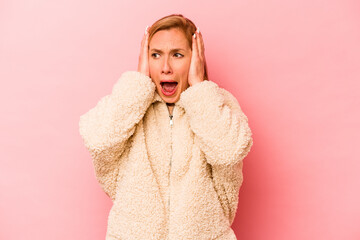 Young caucasian woman isolated on pink background covering ears with hands trying not to hear too loud sound.