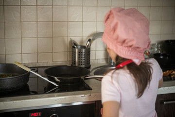 Dziewczynka stoi przed kuchenką i gotuje na patelni  - obrazy, fototapety, plakaty