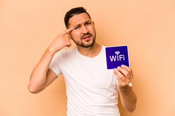 Hispanic man holding wifi placard isolated on beige background showing a disappointment gesture with forefinger.