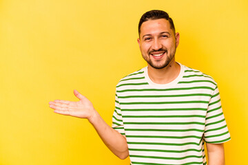 Young hispanic man isolated on yellow background showing a copy space on a palm and holding another hand on waist.
