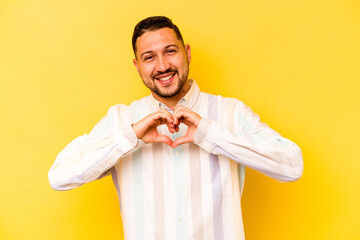 Young hispanic man isolated on yellow background smiling and showing a heart shape with hands.