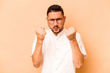 Fototapeta na wymiar Young hispanic man isolated on beige background showing fist to camera, aggressive facial expression.