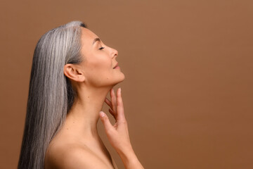 Side view at enchanting topless middle aged Asian woman isolated on brown background, charming korean lady with grey hair stands in profile and touching chin gently. Skin care and self love concept