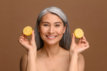 Cheerful attractive middle-aged Asian woman holding lemon near face isolated on bronze background, headshot of eastern female using citric acid for body care and anti aging treatment