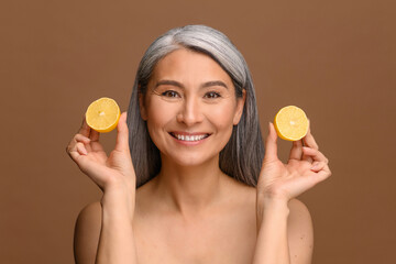 Cheerful attractive middle-aged Asian woman holding lemon near face isolated on bronze background,...