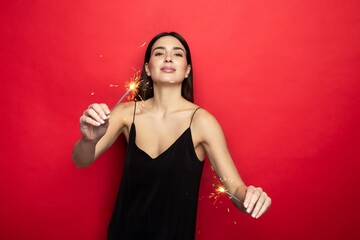 Smiling young woman in a dress holding sparklers on a red background