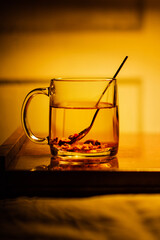 A glass mug with herbal tea on a bedside table, spoon inside the mug. Warm, tungsten light.