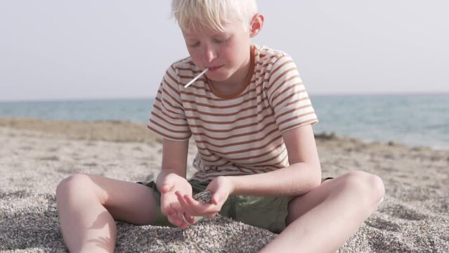 A blond boy plays with sand on the beach by the sea in the summer.