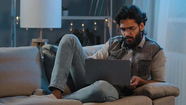 Freelancer guy Arab Indian man bearded male user wearing glasses sitting on sofa at night late time with laptop reading news online thinking doubts job problems pondering solution looking at screen