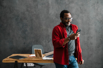 Black man in eyeglasses using cellphone while standing by desk