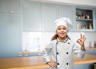 cooking, culinary and profession concept - happy smiling little girl in chef's toque and jacket showing ok gesture over home kitchen background