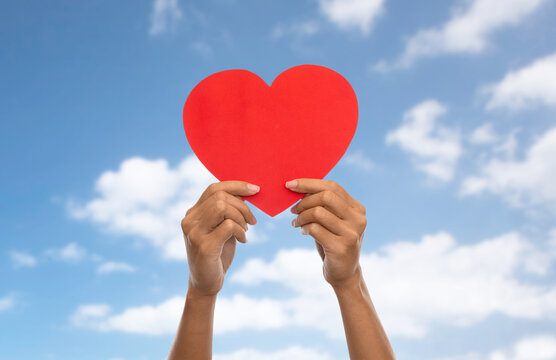 charity, love and health concept - close up of hands holding red heart over blue sky and clouds background