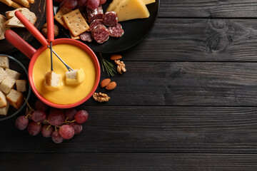 Pot of tasty cheese fondue and snacks on black wooden table, flat lay. Space for text