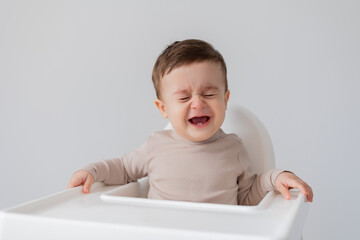 sad upset baby is sitting in a white high chair for feeding. little boy is crying