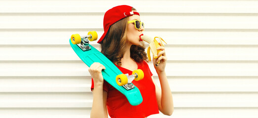 Fashionable portrait of beautiful young woman eating banana with skateboard on white background