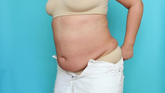 Side view of unrecognizable fat plump overweight woman wearing beige underwear, putting on, adjusting, buttoning up white jeans on blue background. Body positive, obesity, weight loss, liposuction.