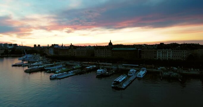 Aerial Backward Shot Of Nautical Vessel Moored In Sea By City Against Cloudy Sky During Sunset - Stockholm, Sweden