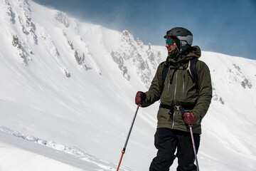 male skier in jacket with goggles and a ski helmet and ski poles stands against the backdrop of snow-capped mountains