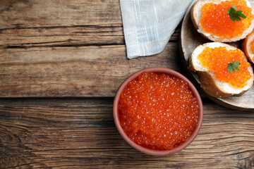 Bowl with delicious red caviar and tasty sandwiches on wooden table, flat lay. Space for text