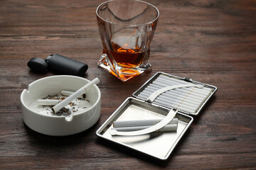 Open case with tobacco filter cigarettes, ashtray, lighter and alcohol drink on wooden table
