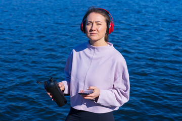 A young athletic woman listens to music and drinks water during breaks from outdoor training