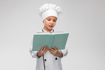 cooking, culinary and profession concept - happy smiling little girl in chef's toque and jacket reading cook book over grey background